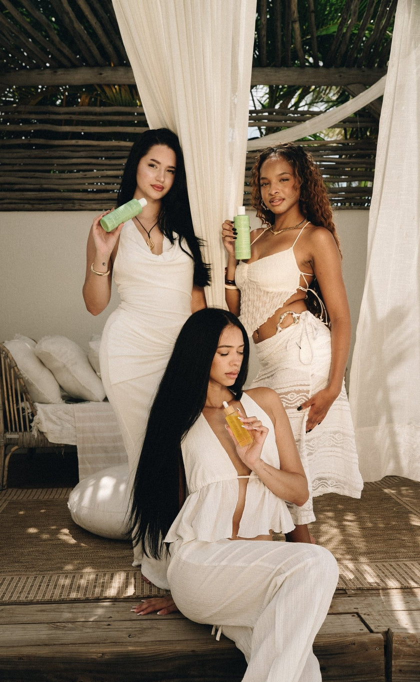 Three women in white holding Mimane Glow shampoo, conditioner, and hair oil under a draped canopy